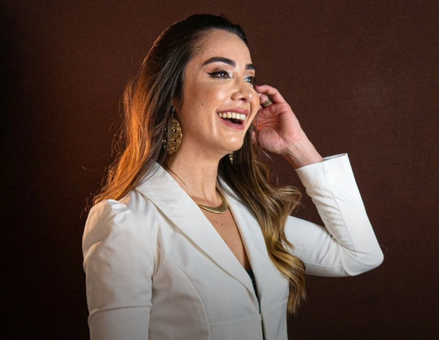 Dra. Mariana Abdalla sorrindo, de blazer branco, diante de um fundo marrom escuro.